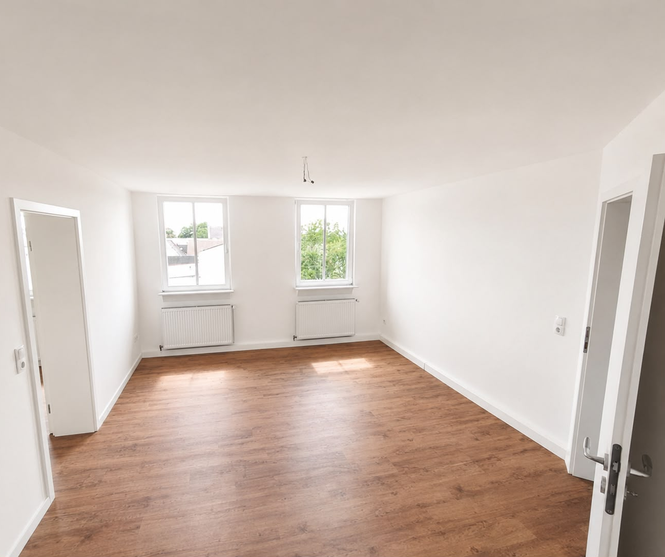 Heller Wohnraum mit zwei Fenstern, weißen Wand- und Deckenflächen sowie Holzfußboden