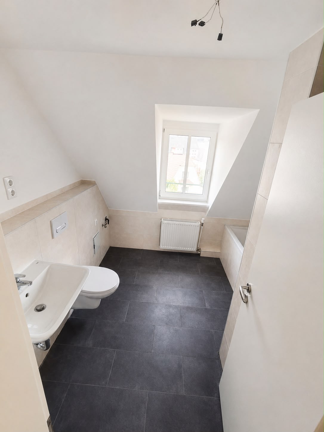 Dachgeschossbad im Hochformat mit Waschbecken und WC links, Fenster und Heizkörper mittig sowie Badewanne rechts